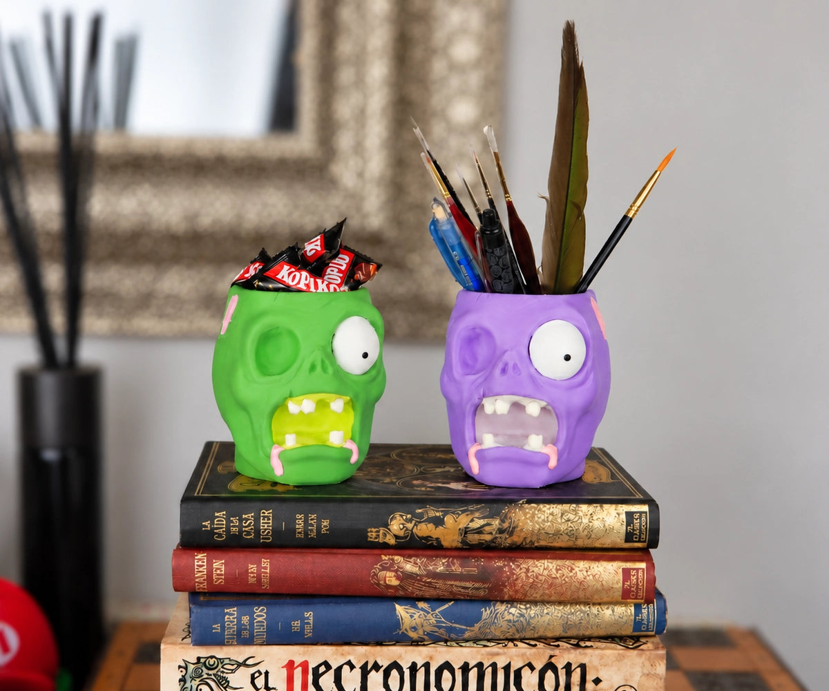 Grüne und lila Zombie-Mehrzweckschalen auf einem Stapel Horrorbücher, eine gefüllt mit Halloween-Süßigkeiten und die andere mit Schreibutensilien
