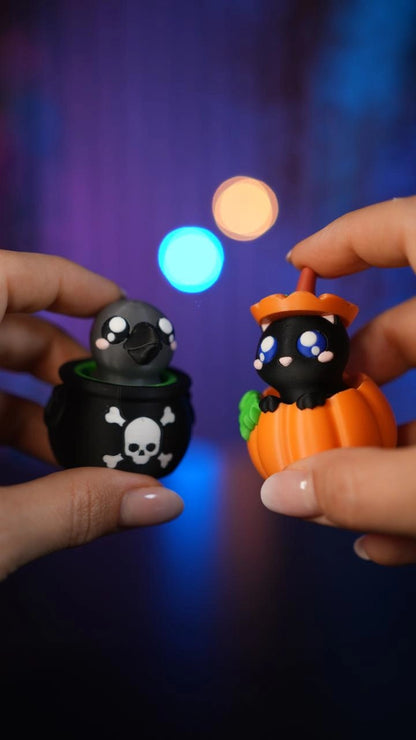 Hände halten zwei Halloween-Schlüsselanhänger-Clicker: einen als Kessel mit Totenkopf und einen in Kürbisform - Kokoroto