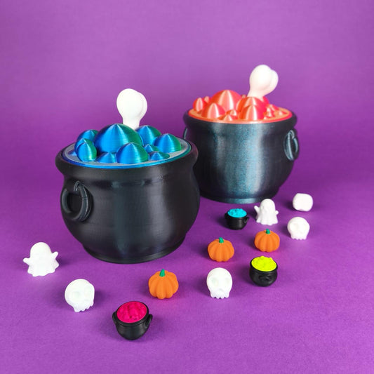 Die beiden Hexenkessel-Süßigkeitenschalen, eine mit kleinen Figuren von Geistern, Kürbissen und Totenköpfen, die andere mit einem sprudelnden pinken Zaubertrank, vor violettem Hintergrund - Kokoroto