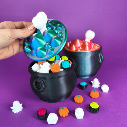 Mano levantando la tapa del caldero de bruja para caramelos, mostrando las figuritas de colores en su interior - Kokoroto